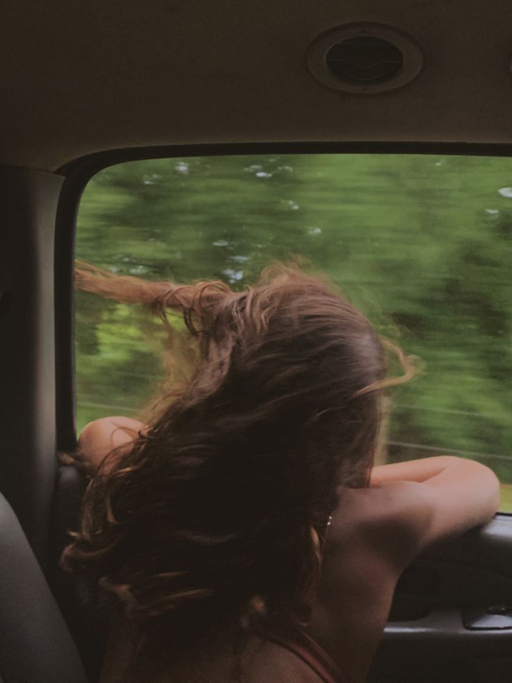 Person leaning out of a car window, hair blowing in the wind with green trees passing by.