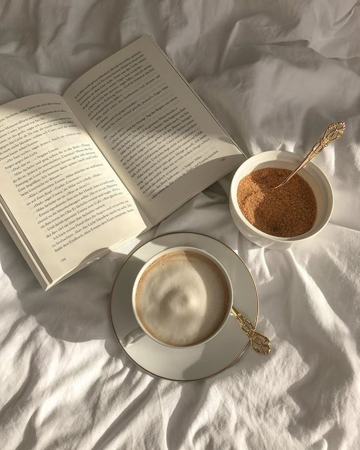 An open book, a cup of coffee and a bowl of sugar on soft white sheets in warm sunlight.